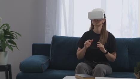 young-woman-is-using-modern-HMD-display-on-head-is-sitting-living-room-moving-hands-controlling-by-virtual-screen-modern-technology-of-virtual-reality